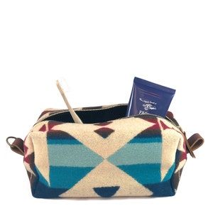 May include: A rectangular toiletry bag featuring a geometric pattern in shades of cream, turquoise, dark blue, and maroon. The bag has a zippered opening, brown leather side tabs, and contains a light-colored toothbrush and a dark blue tube.