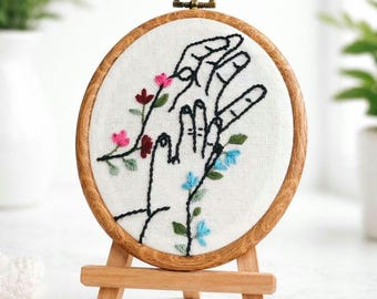 May include: Oval embroidery hoop with a wooden frame, featuring a hand-stitched design of two hands reaching towards each other. The design is accented with delicate floral embroidery in pink and blue. The hoop is displayed on a small wooden easel.