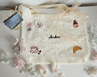 May include: A cream-colored canvas tote bag featuring embroidered designs of a pink rose, a cupcake, a coffee cup with steam, and a croissant. The name Ambar is embroidered in dark thread in the center. A sheer beige ribbon is tied to the handle.