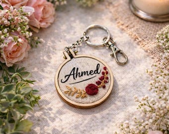 May include: A personalized keychain with the name "Ahmed" embroidered on a circular wooden frame. The design includes floral accents in red and gold, attached to a silver chain and clasp. The background features flowers and a lit candle.