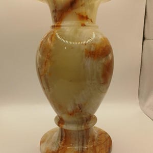 May include: A polished stone vase featuring a smooth, rounded body with a flared rim and a circular pedestal base. The surface displays a translucent, marbled pattern with swirling bands of cream, pale green, and deep amber-orange mineral veins.