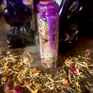 May include: A small glass bottle filled with dried herbs and seeds, sealed with a cork and purple wax. The wax drips down the sides of the bottle. Dried flowers and herbs are scattered around the base of the bottle. Two black raven figurines are in the background.