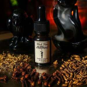 May include: A dark glass bottle of "Black Hene Stillbind" candle dressing oil for binding, 15 ml, with a black dropper. The bottle sits on a silver tray with dried herbs and cloves, near a black incense burner and a black decorative object.