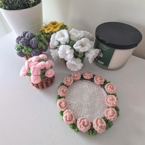 May include: A collection of handmade crochet items including three small potted flower arrangements in purple, yellow, and white, a small pink flower pot, and a circular white coaster decorated with a border of small pink crocheted roses.