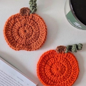Puede incluir: Dos posavasos redondos de ganchillo con forma de calabaza naranja. Cada posavasos presenta un pequeño tallo de ganchillo marrón y un zarcillo de ganchillo verde rizado unido a la parte superior. La textura se crea con filas circulares de hilo.