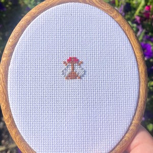 以下が含まれることがあります： 小さな丸い刺繍枠。キノコのクロスステッチのデザイン。キノコは赤と茶色で、白い背景に配置されています。緑の葉と紫の花を背景に、枠が保持されています。