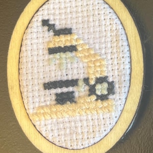 Puede incluir: Un llavero de punto de cruz hecho a mano con un pequeño diseño bordado dentro de un aro de madera. El diseño incluye puntadas negras, amarillas y verde claro sobre un fondo blanco. El llavero tiene un cierre dorado.