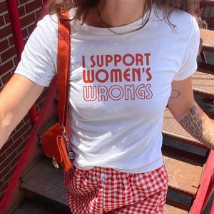 以下が含まれることがあります： 赤い文字で「I SUPPORT WOMEN'S WRONGS」と書かれた白いTシャツ。赤と白のチェック柄のショートパンツ。オレンジ色のショルダーバッグも見える。