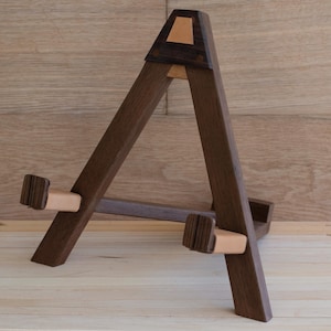 Puede incluir: Un caballete de madera marrón oscuro con un diseño en forma de A. Cuenta con detalles en cuero marrón claro y está diseñado para sostener un lienzo o una obra de arte. El caballete está sobre un fondo de madera clara.