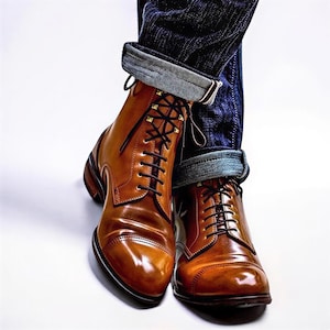 Könnte beinhalten: Ein Paar polierte Schnürstiefel aus hellbraunem Leder mit dunkelbraunen Schnürsenkeln und goldfarbenen Ösen. Die Stiefel haben eine Zehenkappe und werden zu einer dunkelblauen Jeans getragen, die am unteren Saum einen hellblauen Umschlag zeigt.