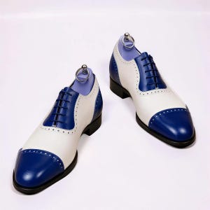 Könnte beinhalten: Ein Paar zweifarbige Oxford-Schuhe mit königsblauer Zehenkappe und Fersenkappe im Kontrast zu weißen Seitenteilen. Diese Schnürschuhe verfügen über dunkelblaue Schnürsenkel, dekorative Brogue-Details an den Nähten und eine schwarze Sohle.