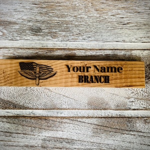 May include: Wooden desk sign with engraved text and an emblem featuring an American flag and wings. The sign reads "Your Name" above "BRANCH" in a bold, sans-serif font. The wood has a natural grain and is set against a rustic background.