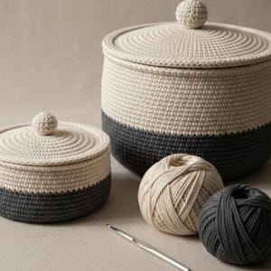 May include: Two-tone crochet storage containers with lids, in cream and charcoal grey. The containers are round, with a textured surface and a small ball on top of the lid. Two balls of yarn and a crochet hook are also visible.