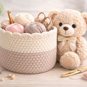 May include: A crocheted basket filled with yarn balls, scissors, and a crochet hook. A plush teddy bear with a bow sits beside the basket. A measuring tape and wooden beads are also on the table. The color palette includes cream, pink, and brown.