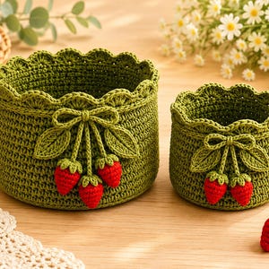 May include: Two round, olive green crocheted baskets with scalloped edges. The larger basket features three small red crocheted strawberries with green leaves and a bow, while the smaller basket has two strawberries with leaves and a bow.