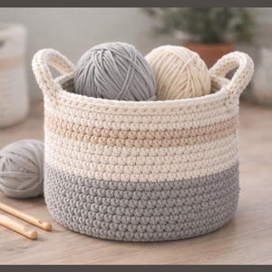 May include: A round, striped crochet basket in grey, beige and cream, with two handles. The basket contains two balls of yarn, one grey and one cream. Two wooden crochet hooks are next to the basket. A second basket is in the background.