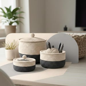 May include: Three woven storage containers with lids and an open container. The containers have a cream-coloured top and a dark grey base. The containers are on a light-coloured table. Pens and a ball of yarn are visible.