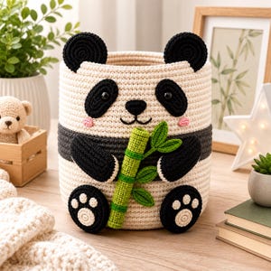 May include: A cylindrical crochet storage basket designed to look like a panda. The basket is cream-colored with black ears, eye patches, arms, and feet. It features a small pink blush on the cheeks and holds a bright green crocheted bamboo stalk.