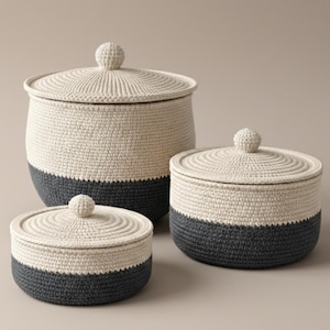 May include: Three hand-crocheted storage baskets with lids. The baskets are cream-coloured with a dark grey base. Each basket has a round, knob-like handle on the lid. The baskets are different sizes, suitable for various storage needs.