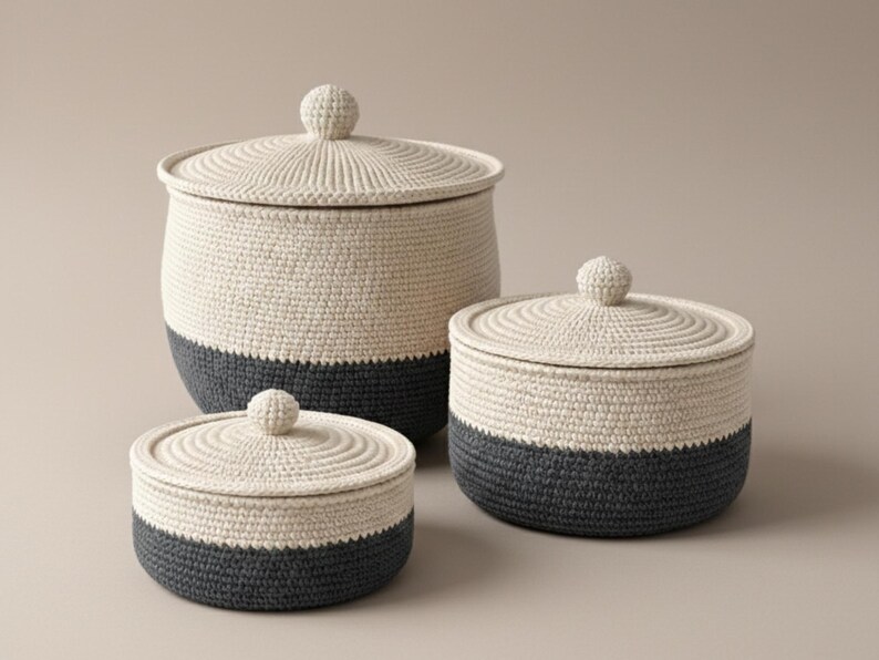 May include: Three hand-crocheted storage baskets with lids. The baskets are cream-coloured with a dark grey base. Each basket has a round, knob-like handle on the lid. The baskets are different sizes, suitable for various storage needs.