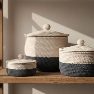 May include: Three handcrafted storage baskets with lids, in varying sizes, sit on a wooden shelf. The baskets are two-toned, with a cream-coloured upper section and a dark grey lower section. Each basket has a round, knob-like handle on the lid.