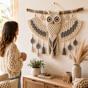May include: A macrame wall hanging shaped like an owl, crafted from cream and dark gray cord. The owl features large circular eyes, spread wings with fringe, and dangling leaf-shaped accents with wooden beads, all suspended from a natural wood branch.
