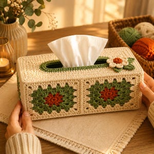 May include: A rectangular, beige crochet tissue box with a green and red floral design. The tissue box has a rectangular opening for tissues to dispense. A decorative flower is on the side. The tissue box is on a beige placemat.
