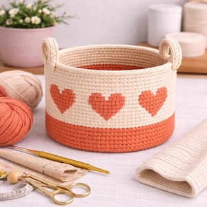 May include: A handmade crochet basket with a cream-colored body and a band of orange at the base. Three orange hearts are evenly spaced around the cream-colored section. The basket has two cream-colored handles. The basket is surrounded by yarn and craft supplies.