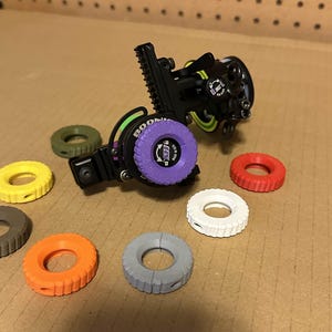 Könnte beinhalten: Ein schwarzes Bogensichtgerät mit einem lila Einstellrad und dem Aufdruck "BOONIE". Mehrere farbige Einstellringe in Gelb, Grün, Orange, Rot, Weiß und Grau sind um das Sichtgerät herum verstreut.