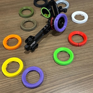 May include: A black archery sight with a green and purple ring, surrounded by various coloured rings in orange, red, yellow, green, purple, white, and black. The sight has a textured surface for precision aiming.