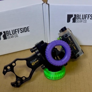May include: A black archery sight with a purple ring and green base. The sight has the text "Imagine Gold" and is positioned in front of two white boxes with the "Bluffside Gear Co." logo.