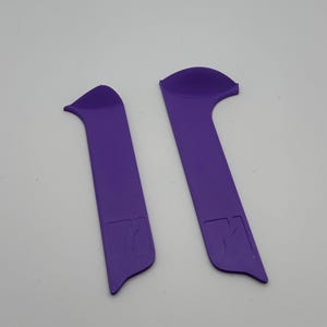 May include: Two purple, elongated, curved objects with a rounded top and a rectangular base. The base of each object has a small, detailed design. The objects are set against a plain, light background.