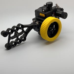 May include: A black archery sight with a yellow adjustment wheel. The sight has a black bracket with the text "TROPHY RIDGE" and multiple adjustment knobs. The sight is designed for precision aiming.