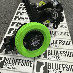 Puede incluir: Un dispositivo mecánico negro y verde con una rueda verde brillante. El dispositivo está etiquetado como "BLUFFSIDE GEAR CO." y "HSS". El fondo presenta múltiples etiquetas blancas y negras con el texto "BLUFFSIDE GEAR CO."