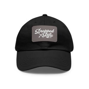 Könnte beinhalten: Schwarze Baseballkappe mit gebogenem Schirm. Die Kappe hat einen grauen rechteckigen Aufnäher mit dem weißen Text "Dropped Rifle Co." in einer stilisierten Schriftart. Der Hut ist aus weichem Material und hat einen klassischen, lässigen Stil.