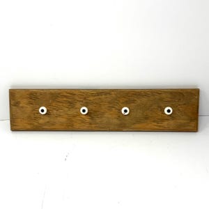 May include: A rectangular wooden wall rack featuring a natural brown wood grain finish. The board is mounted with four evenly spaced, small, round white knobs, each centered with a dark black dot, providing a functional storage solution.