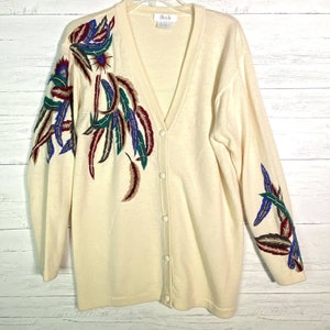 May include: Cream-colored cardigan sweater with a V-neck and button closure. The sweater features embroidered feather designs in shades of burgundy, teal, blue, and brown on the front and sleeves. The sweater has long sleeves and a relaxed fit.