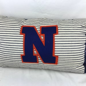 May include: A rectangular throw pillow featuring a white and navy blue pinstriped pattern. The center of the pillow displays a large, dark blue letter N with a thick orange border, and the edges of the pillow are finished with dark blue fabric.