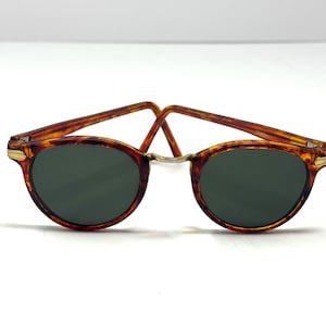 May include: Oval-shaped sunglasses with a tortoiseshell pattern frame and dark green lenses. The bridge and hinges are gold-colored. The sunglasses are set against a white background.