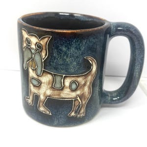 May include: A ceramic mug with a blue glaze and a brown rim. The mug features a whimsical illustration of a dog in brown and cream colors. The handle is a matching blue, and the mug is suitable for hot or cold beverages.