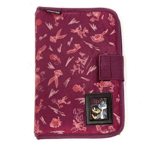 May include: A purple zippered notebook with a repeating pattern of the cartoon characters Wile E. Coyote and Road Runner. The notebook has a black patch with the characters on it and the text "BULLY'S".