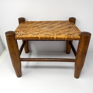 May include: A small wooden stool featuring four thick, cylindrical legs connected by horizontal support bars. The seat is constructed from a woven light brown reed or cane material, showcasing a distinct herringbone pattern across the top surface.