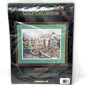 May include: The Gold Collection Christmas Cove counted cross stitch kit by Dimensions in a black plastic package. The kit features a winter village scene with a boat on water, snow-covered buildings, and a Christmas tree. Finished size is 18 by 14 inches.