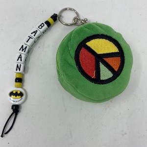 May include: A green plush coin purse with a peace sign design in yellow, red, and green. The purse is attached to a keychain with a black string and a white bead chain that spells out "BATMAN" with yellow and black beads.