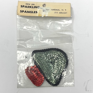 May include: A package of sew-on sparkling spangles. The package contains two sequined appliques: a large, green sequined shape with a black border and a smaller, red sequined shape. The package has a label with the text "SEW ON SPARKLING SPANGLES".