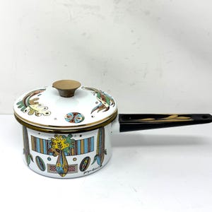 May include: White enamel saucepan with a lid, featuring colorful, stylized illustrations. The pot has a black handle with gold accents and a gold rim. The design includes various shapes and patterns, with the text "georges lancel" visible.