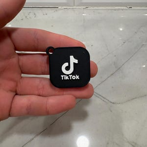 May include: A black square keychain with the TikTok logo in white. The logo features a white musical note and the word "TikTok" in white text. The keychain has a small hole at the top for attachment.