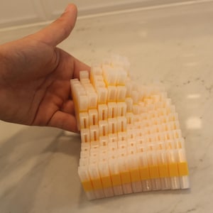 May include: A stack of small, rectangular, off-white plastic pieces with yellow bands. The pieces are arranged in a layered, stepped pattern. The object is held by a hand against a light-colored countertop.