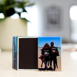 Puede incluir: Una pila de imanes rectangulares. Un imán muestra una foto de dos personas en una playa, con camisetas negras con un logotipo. Los otros imanes son negros. La dirección del sitio web www.pixelstorymagnets.com.au está impresa en el lateral.