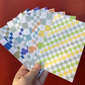 Puede incluir: Una mano sostiene un abanico de hojas de papel rectangulares con un patr&oacute;n de cuadros. Las hojas muestran varias combinaciones de colores, incluyendo azul, verde, amarillo y naranja, dispuestas en un dise&ntilde;o de cuadr&iacute;cula. El fondo es rojo liso.